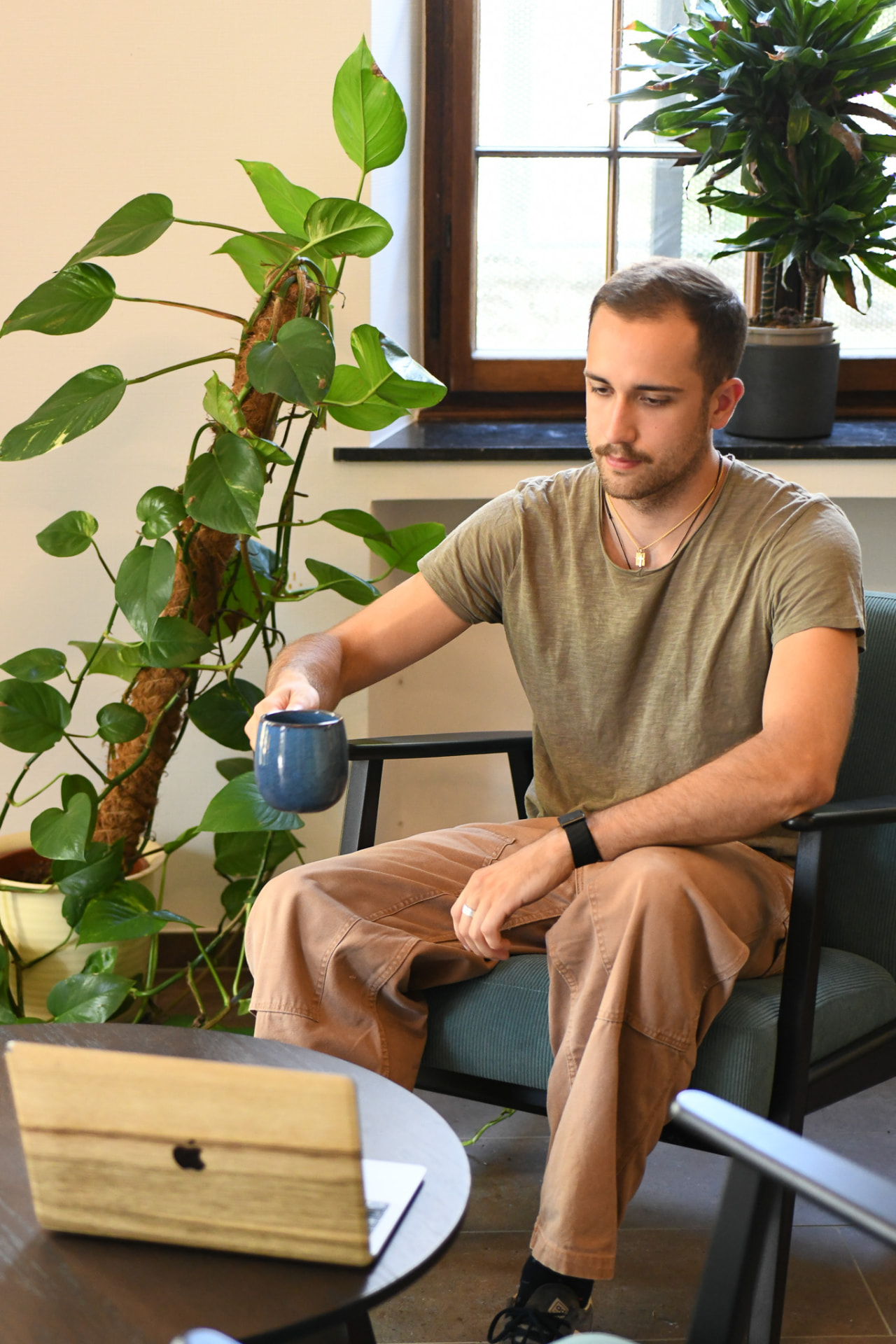 A man working on a laptop in an armchair with a mug, surrounded by plants