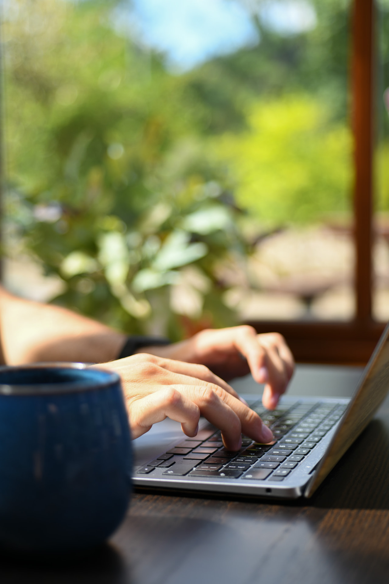Hands typing on a laptop at a wooden table with a blue mug